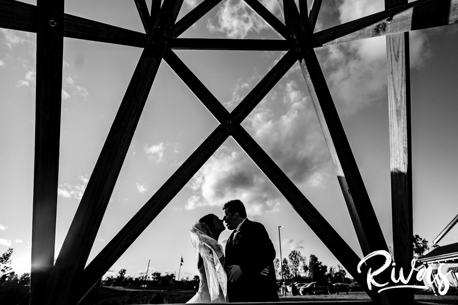 A black and white portrait of a bride and groom leaning in to share a kiss as they stand underneath the legs of a windmill on their fall wedding day at Piazza Messina. 