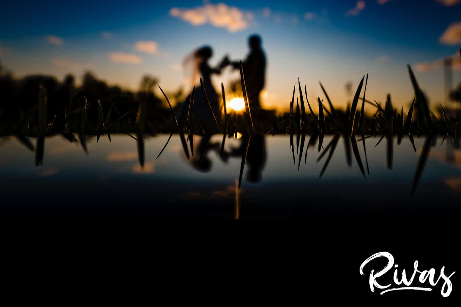 A silhouette of a bride and groom holding hands as the bride's veil blows in the air at sunset, with their reflection visible in the botttom of the frame during their fall wedding day at Piazza Messina in Cottleville, MO.