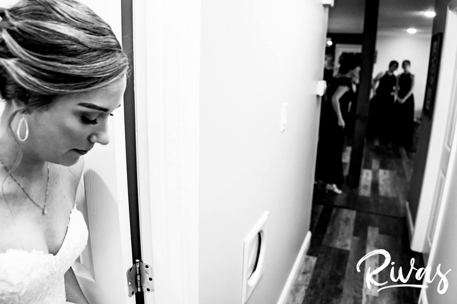 A candid black and white picture of a bride exiting the room where she got dressed to see her bridesmaids for the first time on the morning of her wedding. 