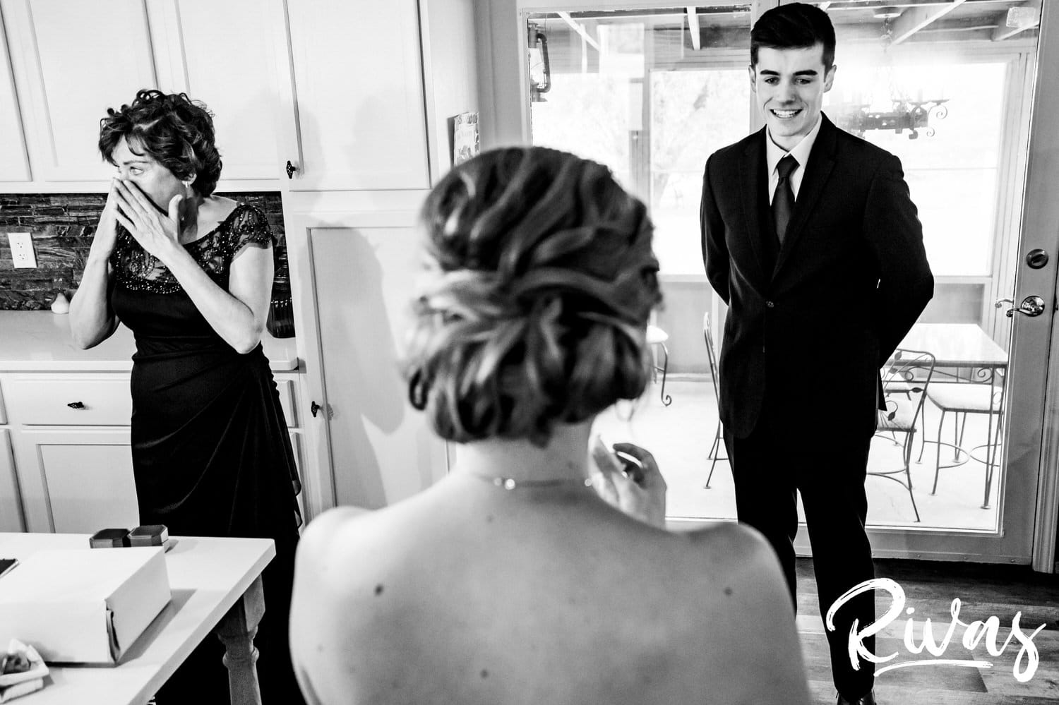 A black and white picture taken over the shoulder of a bride as she sees her younger brother for the first time on her wedding day as her mom cries at the side of the frame. 