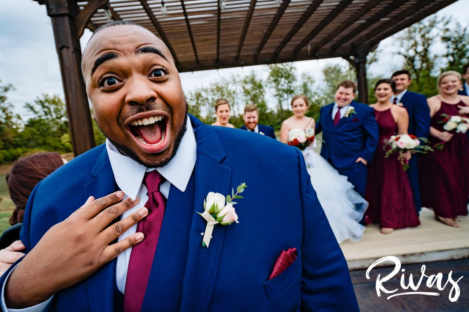 A candid, close-up picture of a groomsmen dramatically doing a "who, me?" gesture as the rest of the wedding party laughs hysterically in the background. 