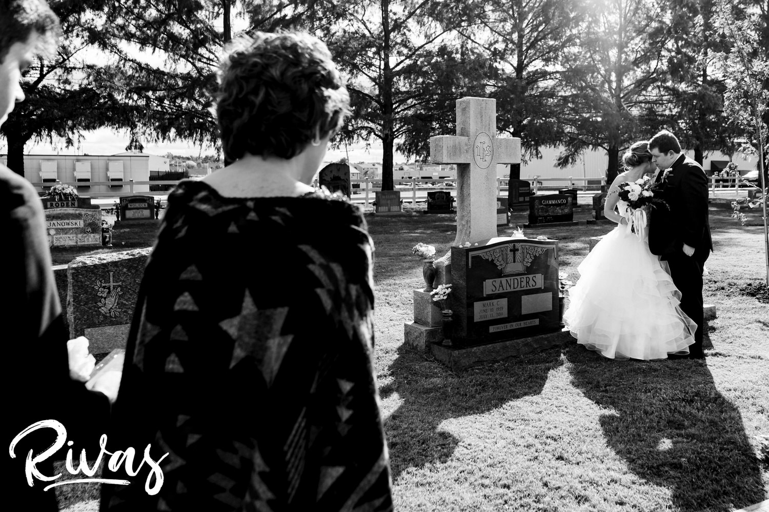 A solemn, black and white picture taken over the shoulder of a bride's mom of a bride and her fiance standing beside her dad's gravestone on a fall wedding day in St. Louis. 