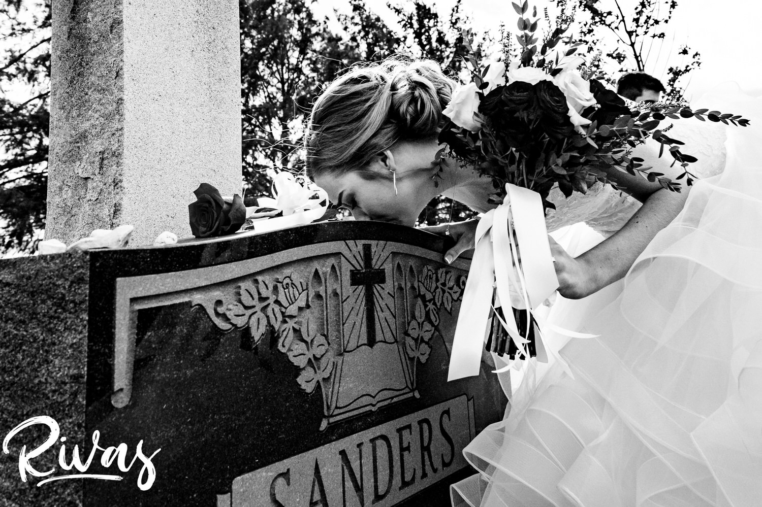 A candid black and white picture of a bride leaning in to kiss the top of her dad's gravestone on her fall wedding day. 