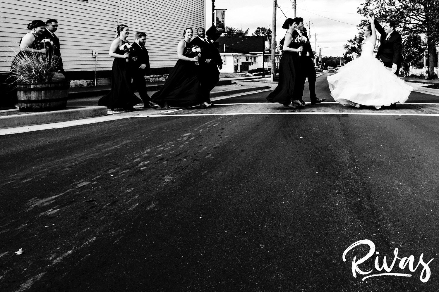 A black and white picture of a bride, groom, and their wedding party lining arms crossing the street in downtown Cottleville, MO on a fall wedding day. 