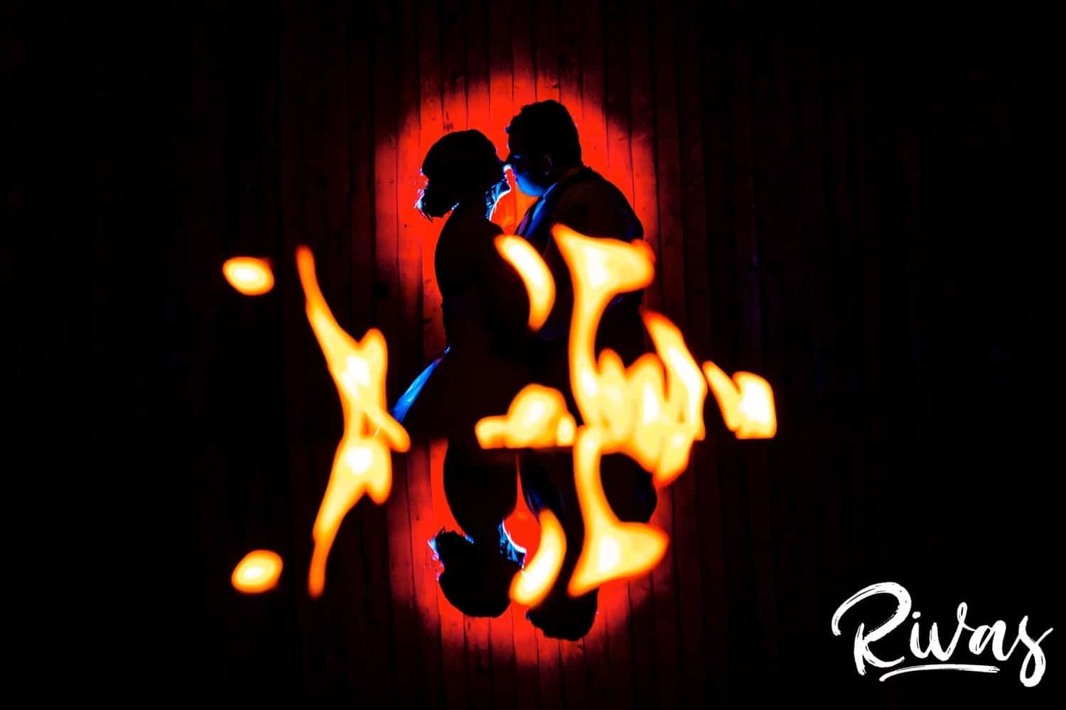 A vibrant silhouetted picture of a bride and groom learning in to share a kiss taken through a fire, with the reflection of both the fire and the couple visible at the bottom of the frame. 