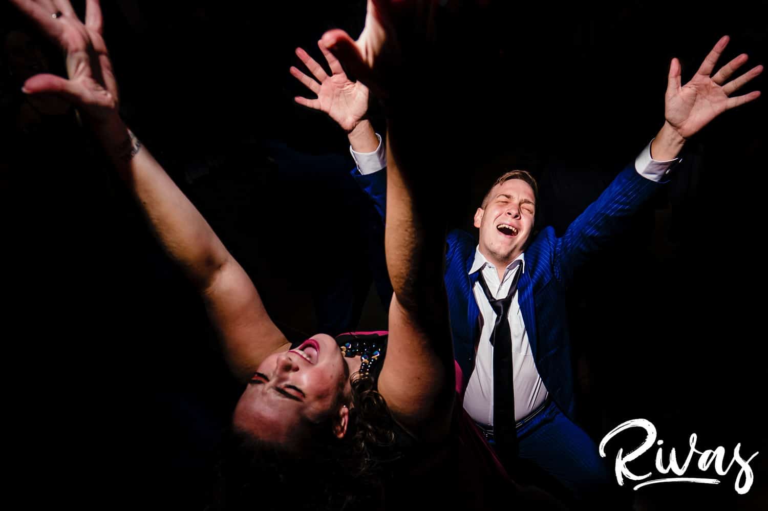 A colorful picture taken from above of two wedding guests throwing their arms up in the air as they dance during a wedding reception. 