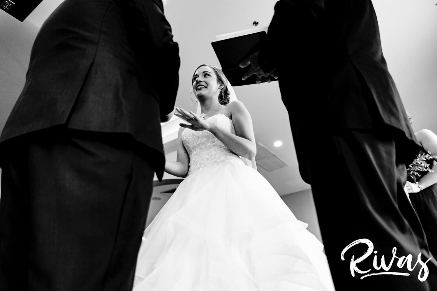 A candid black and white picture taken from below of a groom putting a wedding band on a bride's finger during their wedding ceremony at Piazza Messina. 