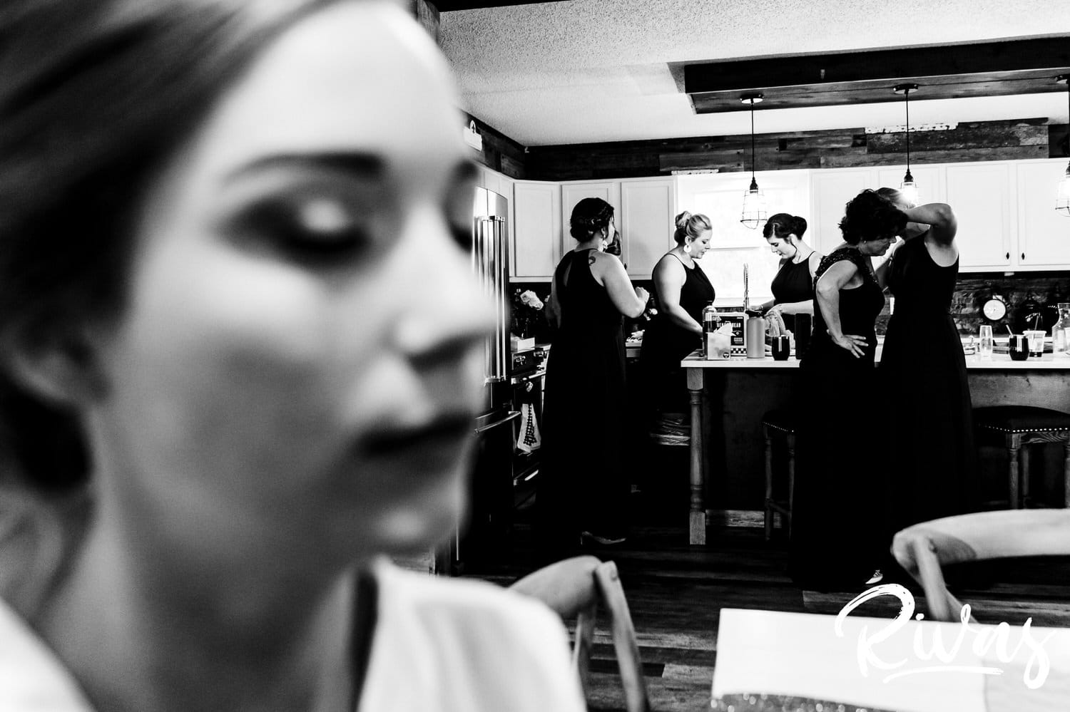 A candid black and white picture of a bride getting her makeup done in the forefront, with her bridesmaids and mom in the background putting on jewelry and zipping dresses on the morning of her fall wedding. 