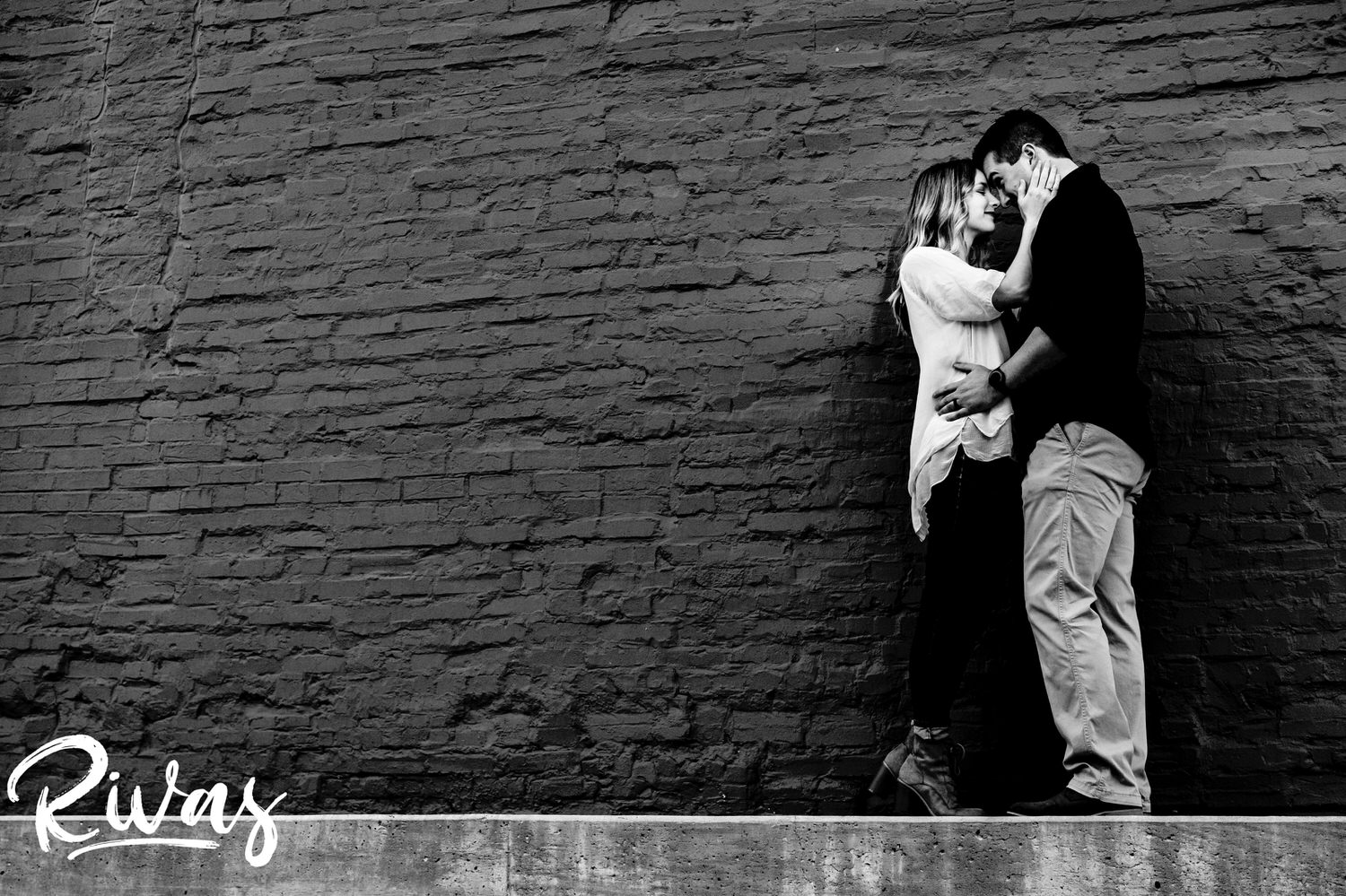 An intimate, full-length, black and white picture of a man and woman sharing a sweet embrace as they stand in front of a painted brick wall during their summer engagement session in Kansas City. 