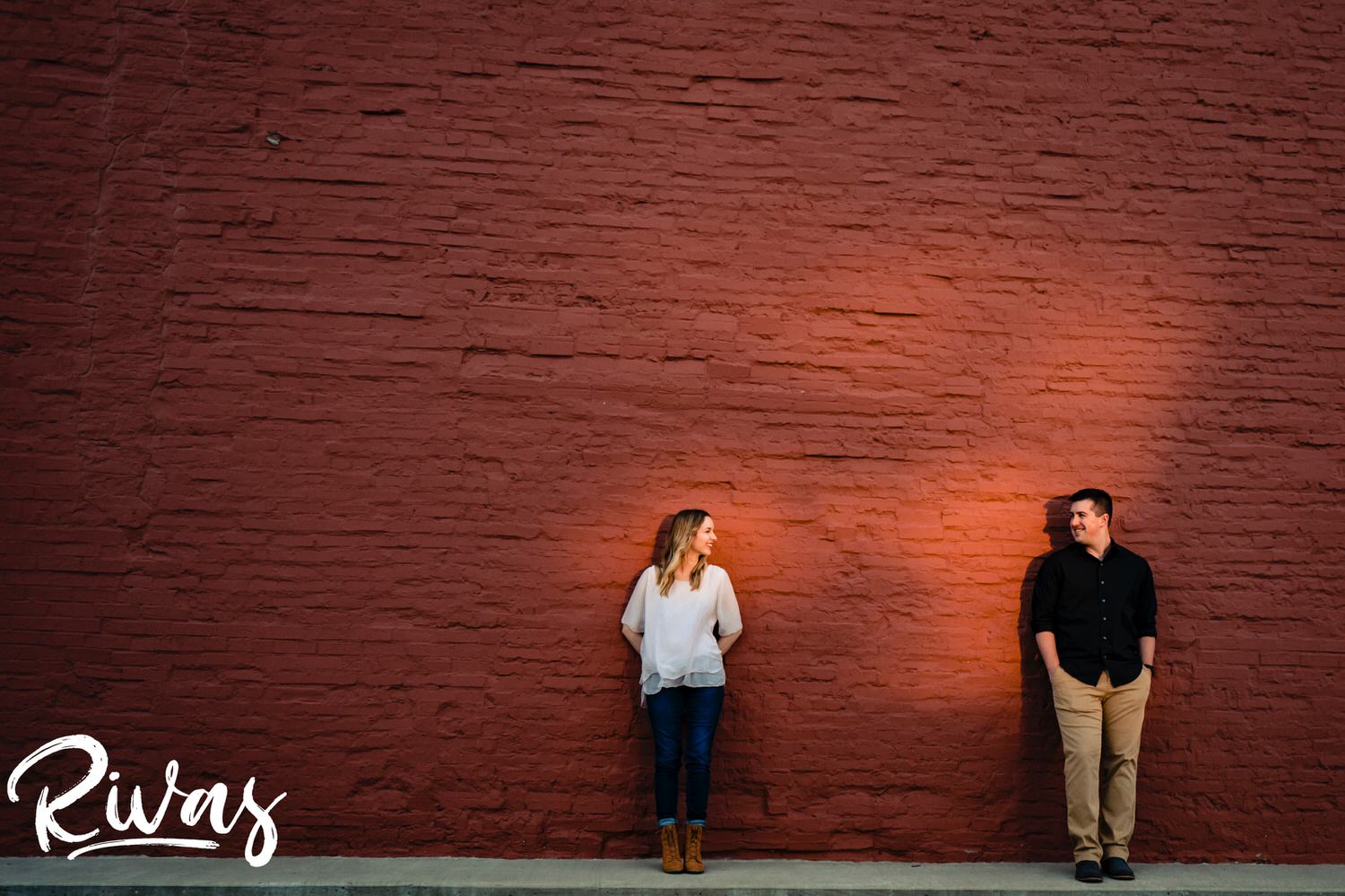 A super-wide, super colorful picture of an engaged couple, each standing in a splash of sunlight, looking up at each other during their summer Kansas City engagement session. 