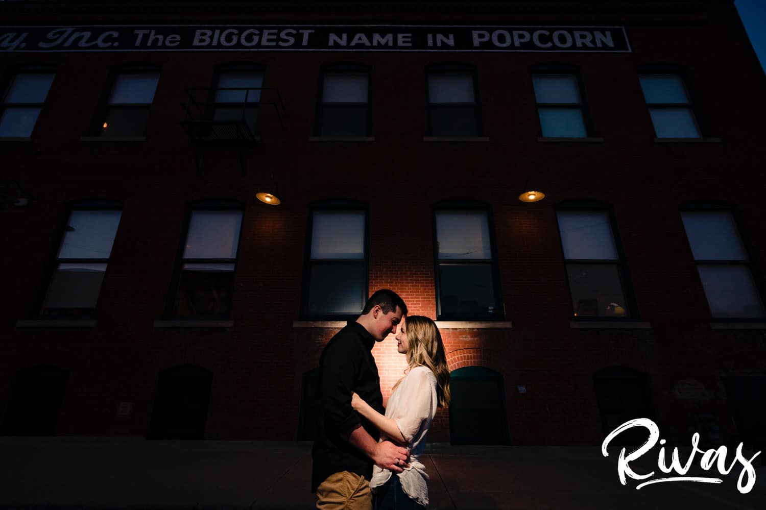 An intimate, wide, rear-lit photo of an engaged couple sharing an embrace as they stand face to face in front of a tall brick building during their City Market summer engagement session. 