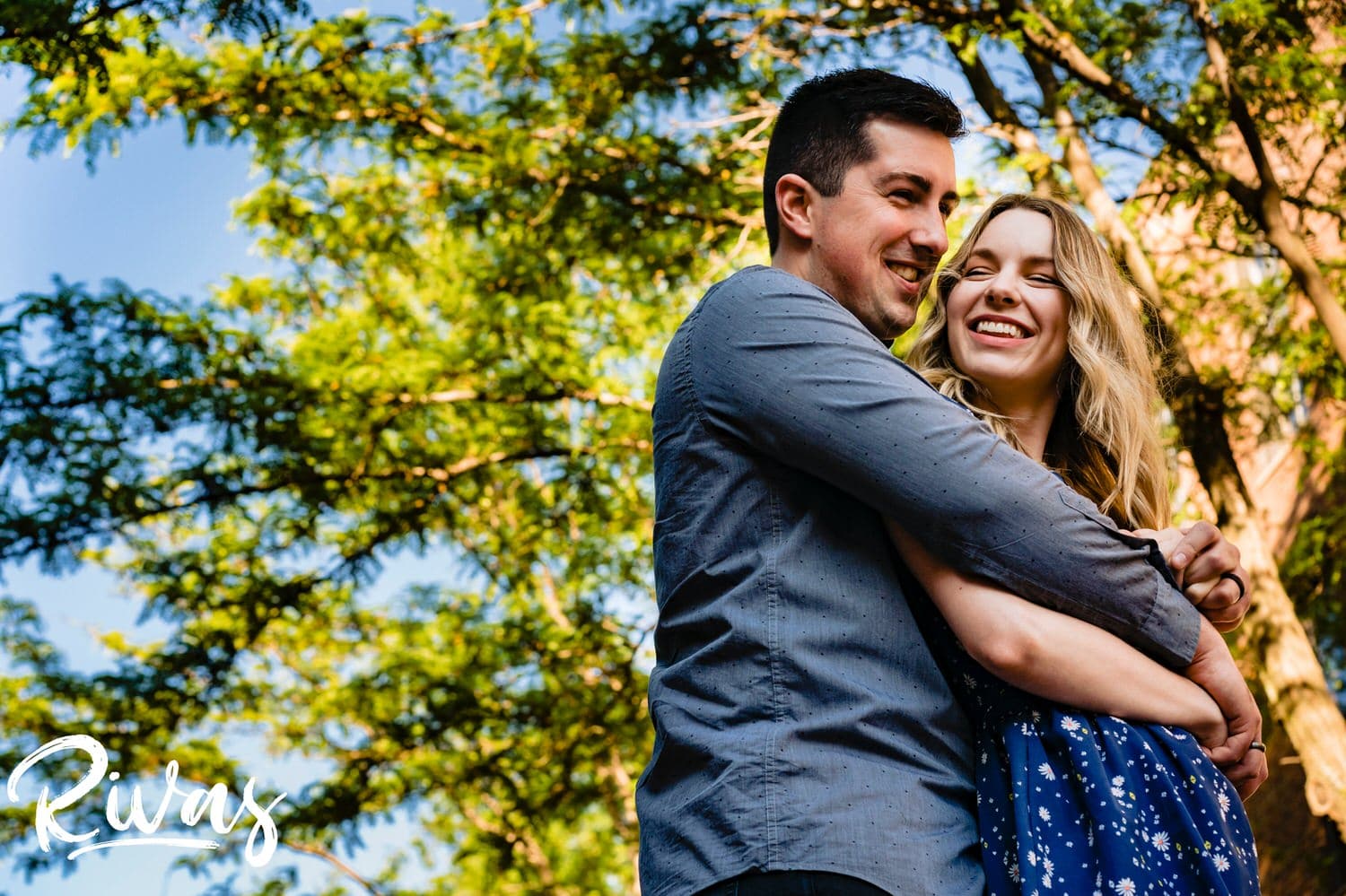 A vibrant, colorful picture of a man hugging his fiance from behind against a colorful canopy of trees in Kansas City's City market during their engagement session. 