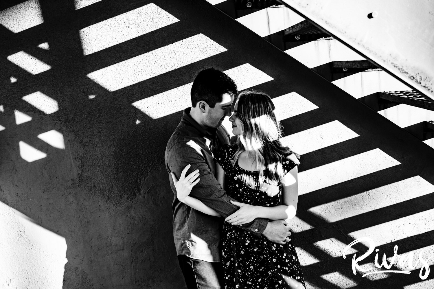 A bold black and white picture of an engaged couple sharing an embrace as they stand on a fire escape, with their shadow visible in the background during their engagement session in Kansas City's City market. 