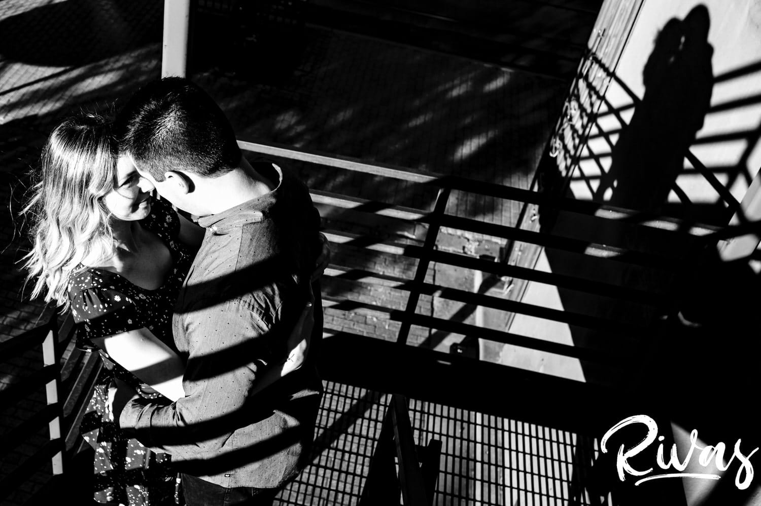 A bold black and white picture of an engaged couple sharing an embrace as they stand on a fire escape, with their shadow visible in the background during their engagement session in Kansas City's City market. 
