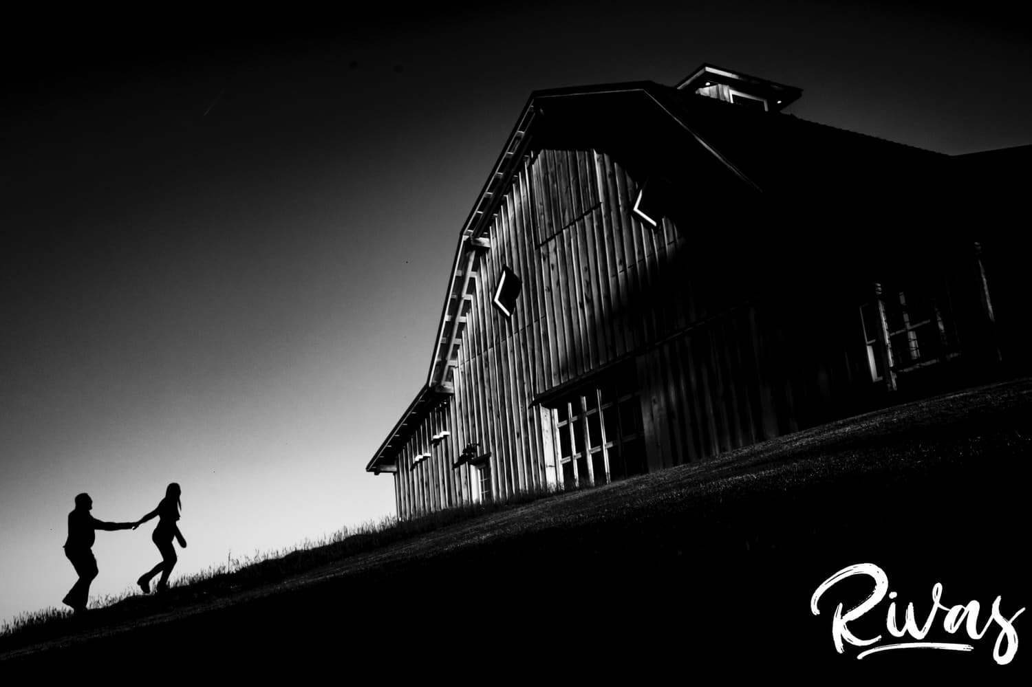 A far-off, black and white picture of a woman leading her fiance up a steep hill towards a giant barn at sunset during the KC engagement session at Schwinn Produce Farm.