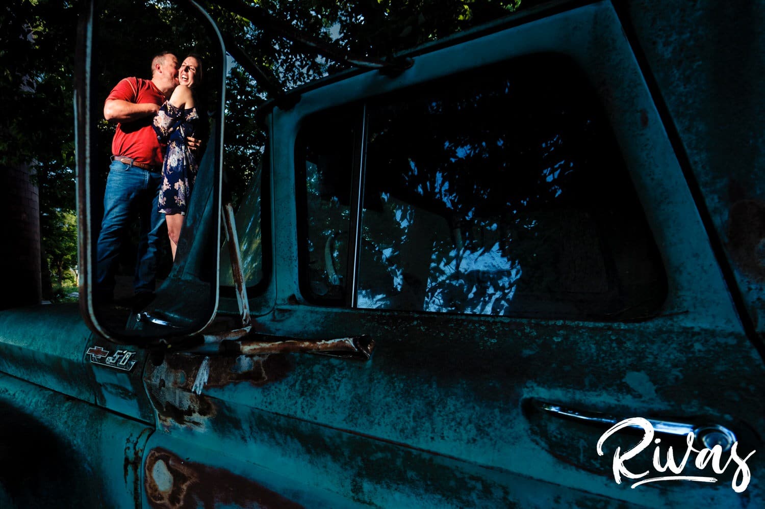 A candid portrait taken in the rear-view mirror of a rusted out blue truck of an engaged couple standing on the flatbed of the truck, laughing together and sharing an embrace during their summer engagement session in Kansas City. 