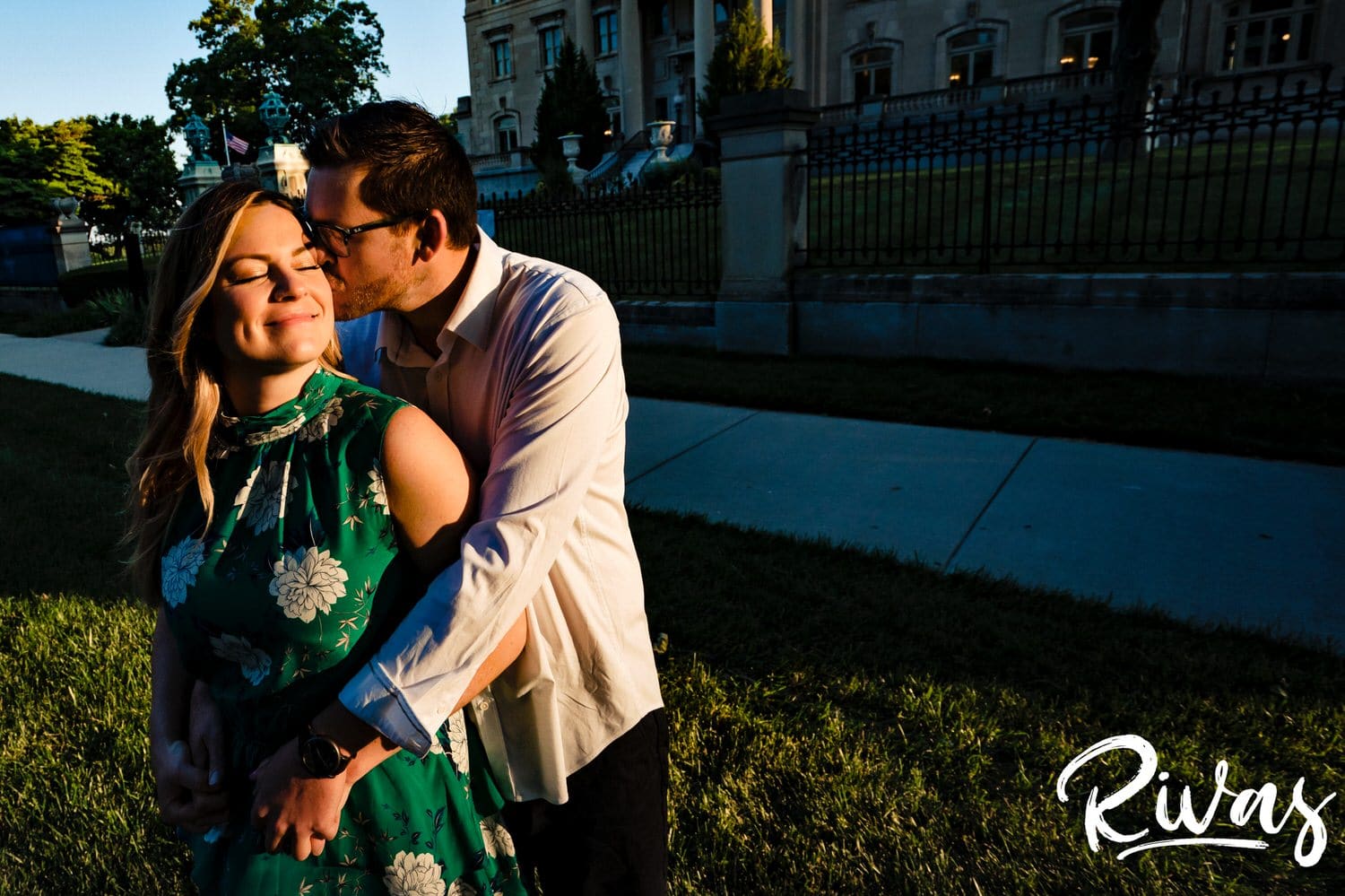 A warm, candid, intimate photo of a man embracing his fiance from behind as he kisses her on the cheek during their summer sunrise engagement session outside the Kansas City Museum.