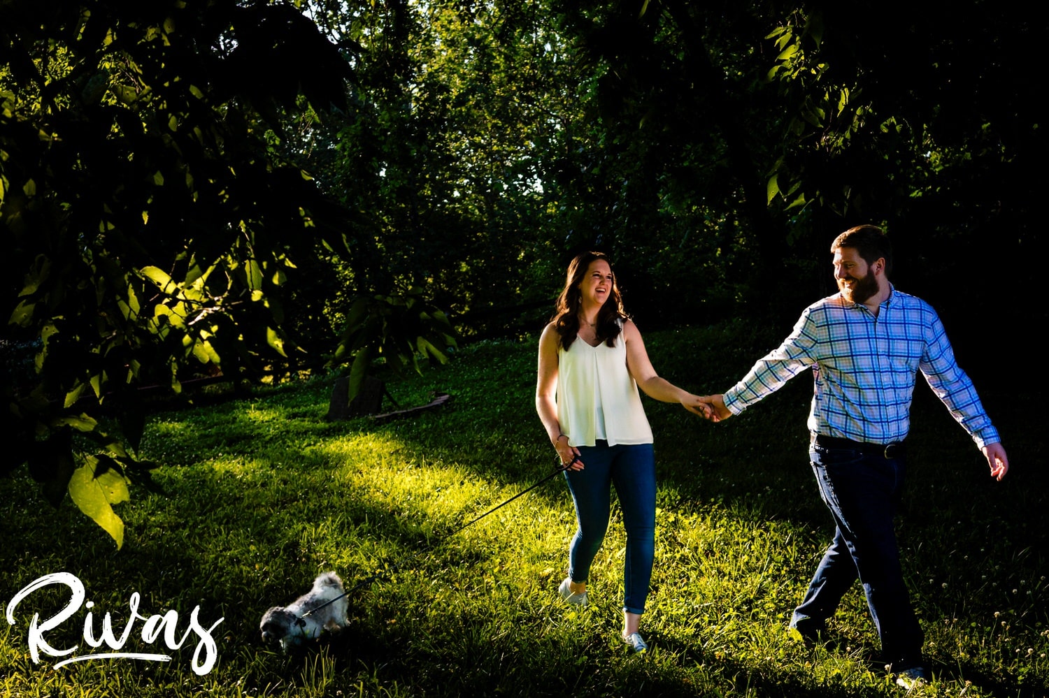 A colorful, greenery filled picture of a man leading his fiance through a park with their dog on a leash during their engagement session at Minor Park in Kansas City. 