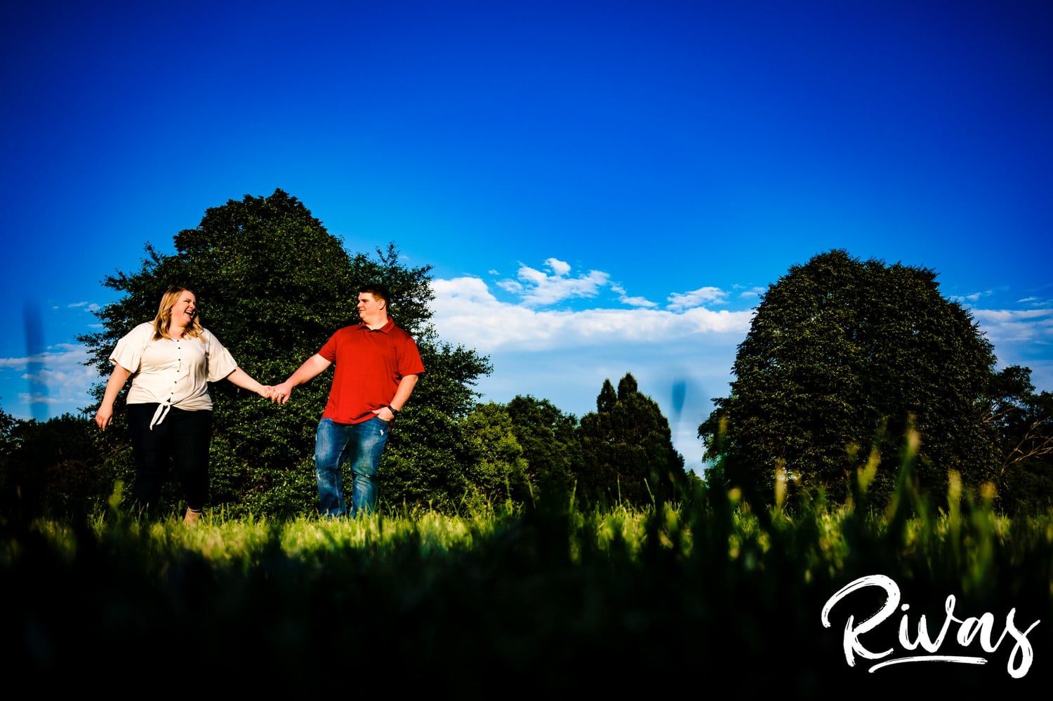 A vibrant, candid picture of an engaged couple holding hands and walking through the grass against a backdrop of green trees and blue skies during their summer engagement session in Kansas City at Loose Park. 