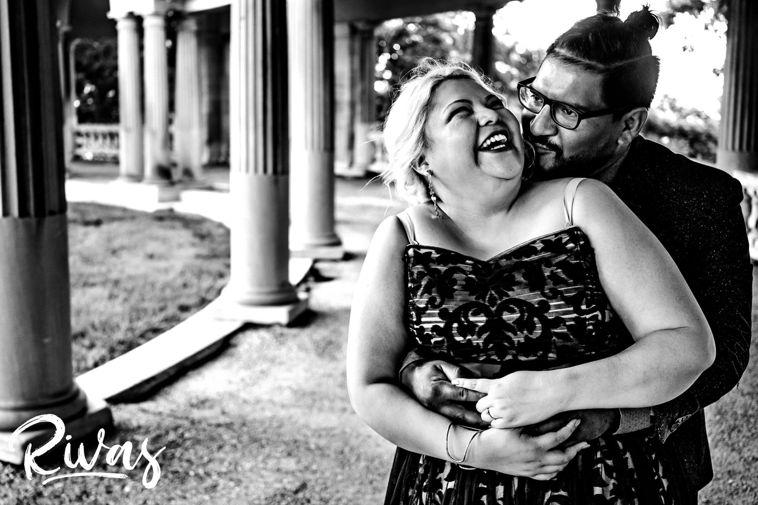 A candid black and white picture of a man in glasses hugging and tickling the blonde woman standing in front of him during their engagement session in Kansas City. 