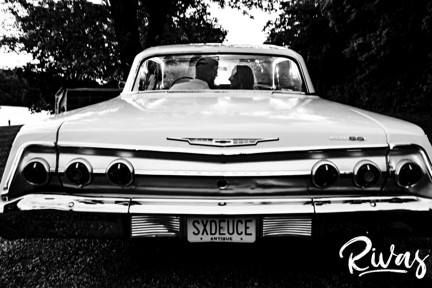 A dramatic black and white picture taken from behind a classic Impala of an engaged couple laughing together and leaning in for a kiss during their summer engagement session in Kansas City. 