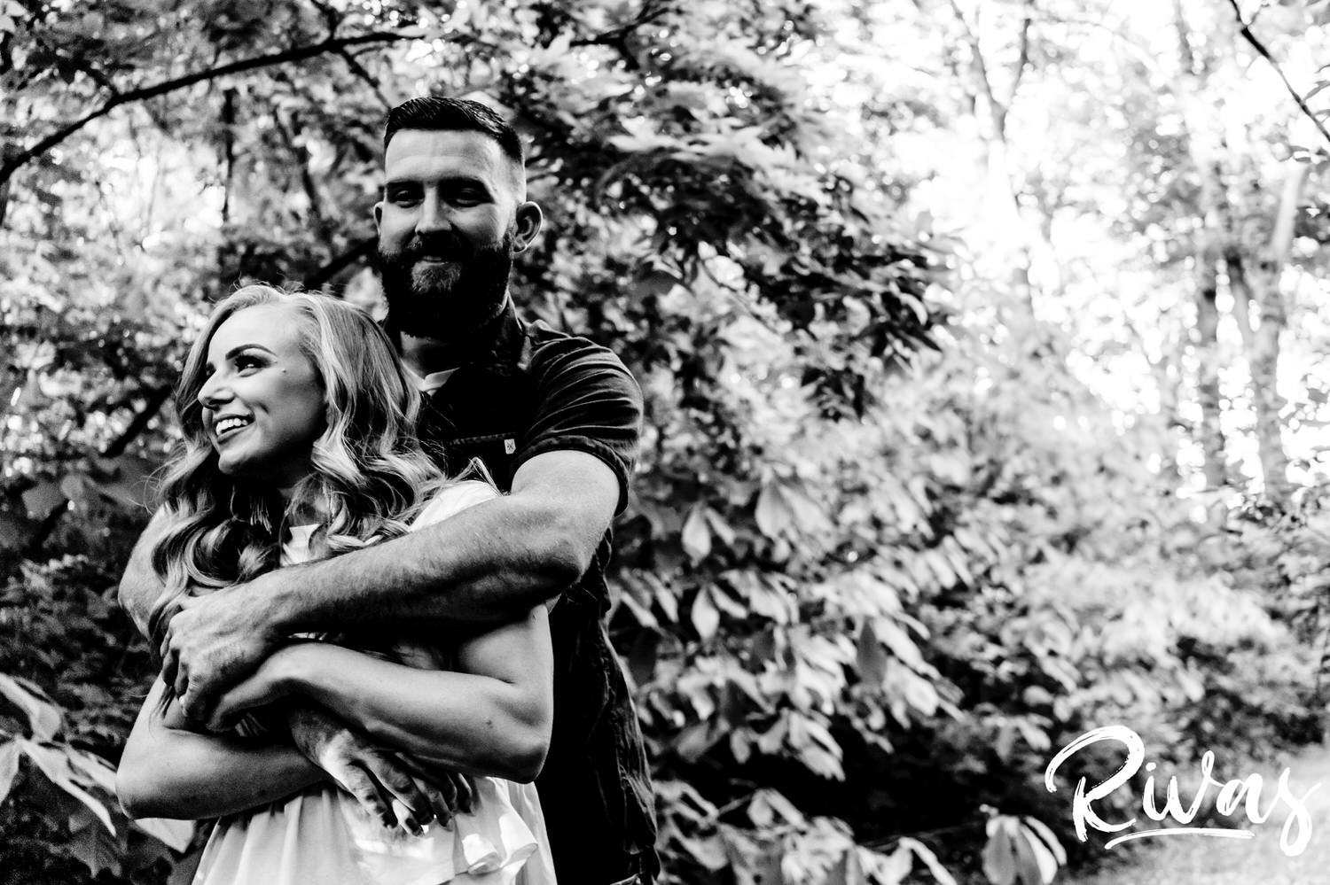 A candid black and white picture of a man with a beard hugging his fiance from behind as she laughs and looks off into the distance during their summer engagement session with their wedding photographer in Kansas City. 