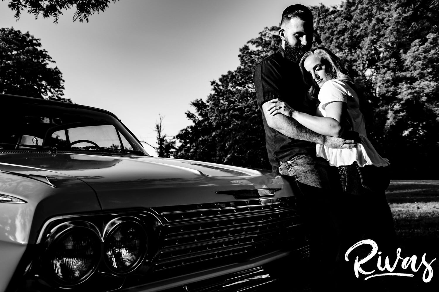 A candid, black and white picture of a couple sharing an intimate embrace as they lean up against a 1969 Impala during their summer engagement session in Kansas City. 