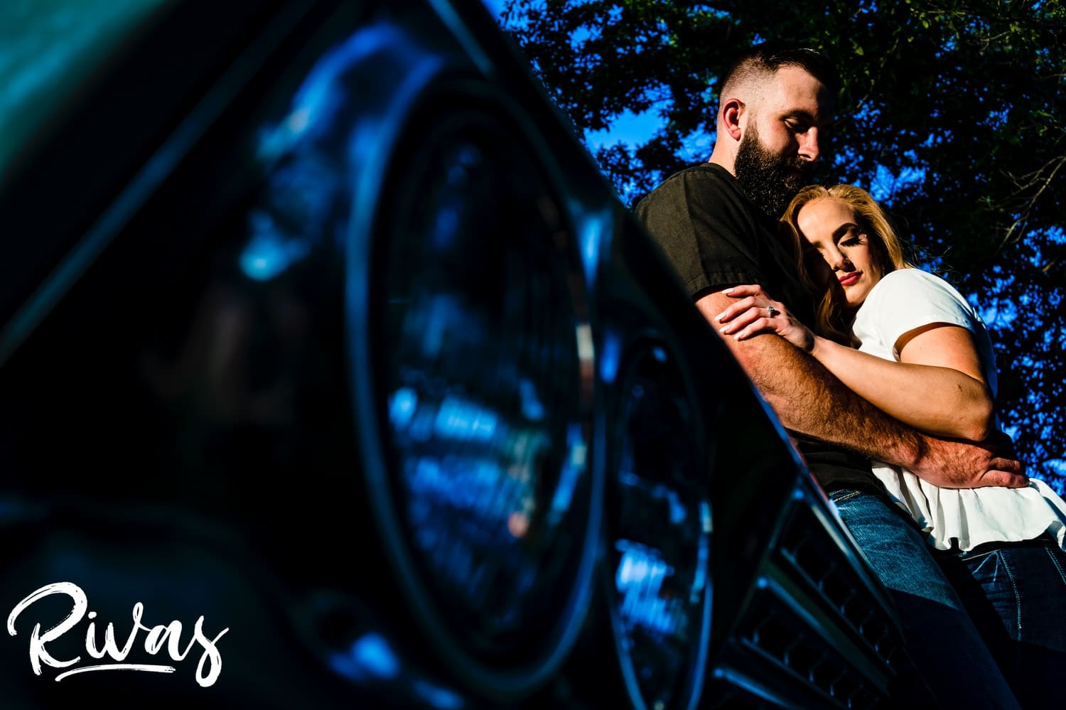 A vibrant portrait of an engaged couple sharing an intimate embrace as they lean up against the fender of an old Impala during their summer engagement session in Kansas City.