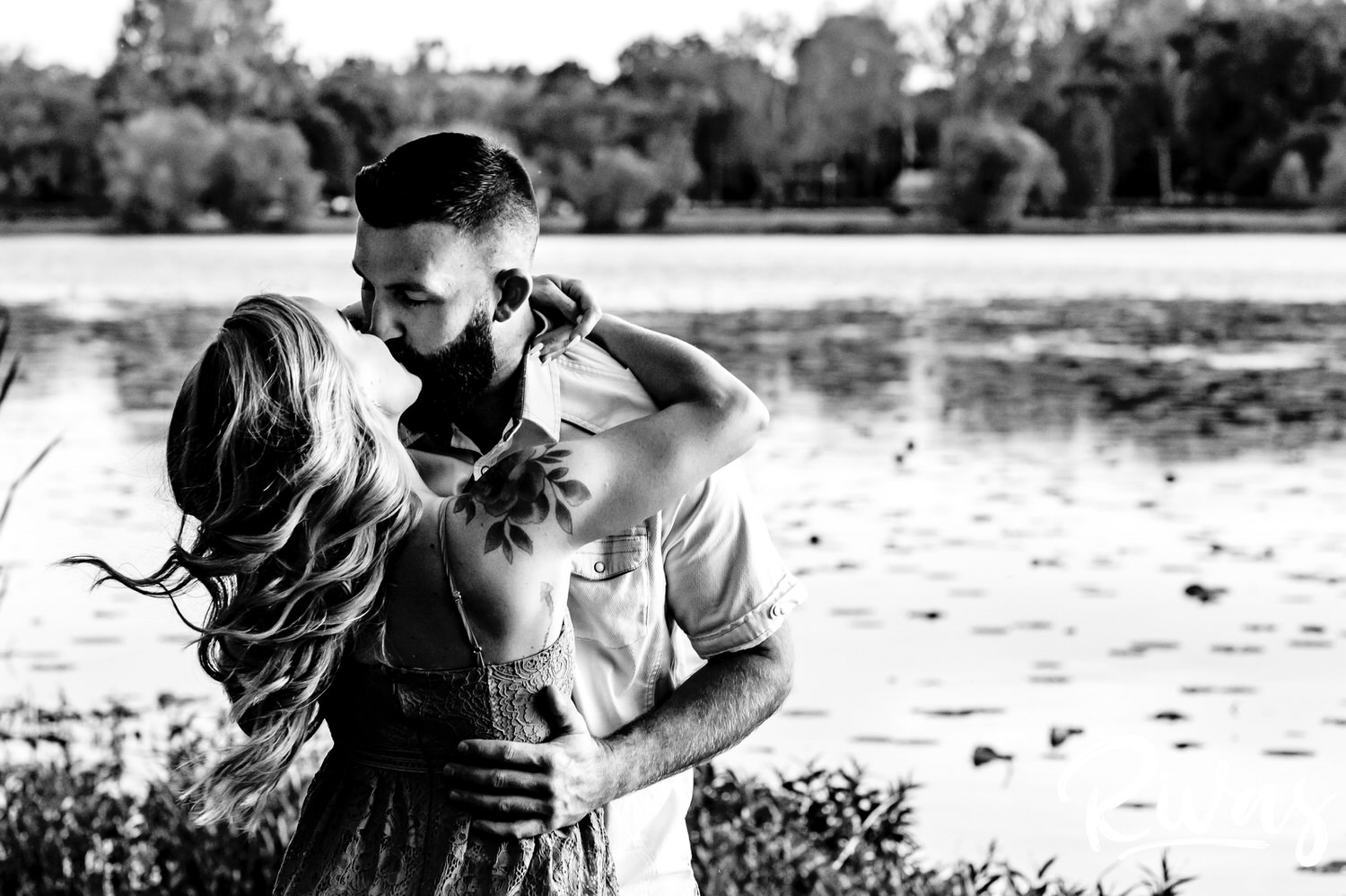 A close-up, intimate picture of a woman leaning in to kiss her fiance as they stand in front of a lake during their summer engagement session in Kansas City. 