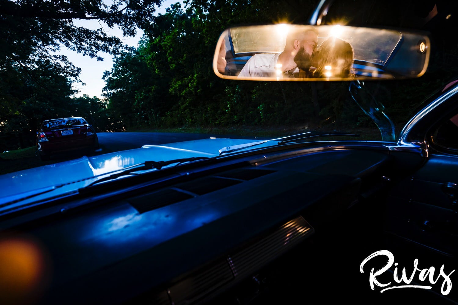 A wide picture taken in the rear-view mirror of an old Impala of an engaged couple leaning in to share a kiss during their summer engagement session in Kansas City. 