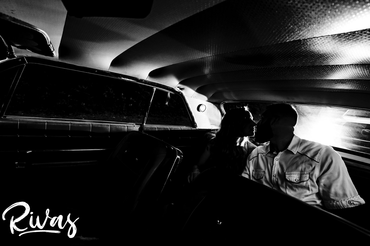 A dramatic black and white picture of an engaged couple sharing a kiss as they sit in the back of a classic Impala during their summer engagement session in Kansas City. 