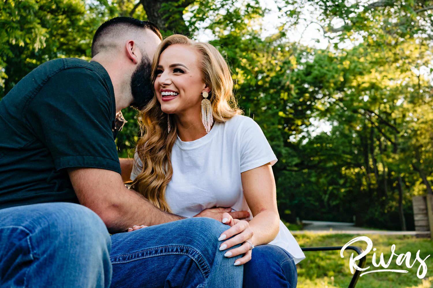 A colorful, candid picture of a man whispering into his fiance's ear as they sit on a bench surrounded by greenery during their summer engagement session with their wedding photographer. 