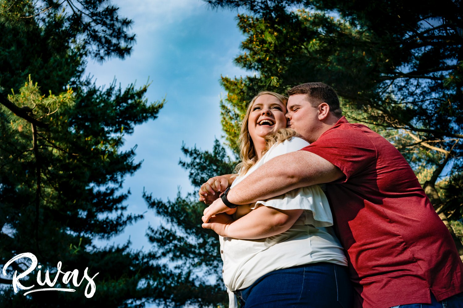 A candid, colorful, tight picture taken from the ground looking up of a man hugging his fiance from behind as she laughs during their engagement photography session in Kansas City at Loose Park. 