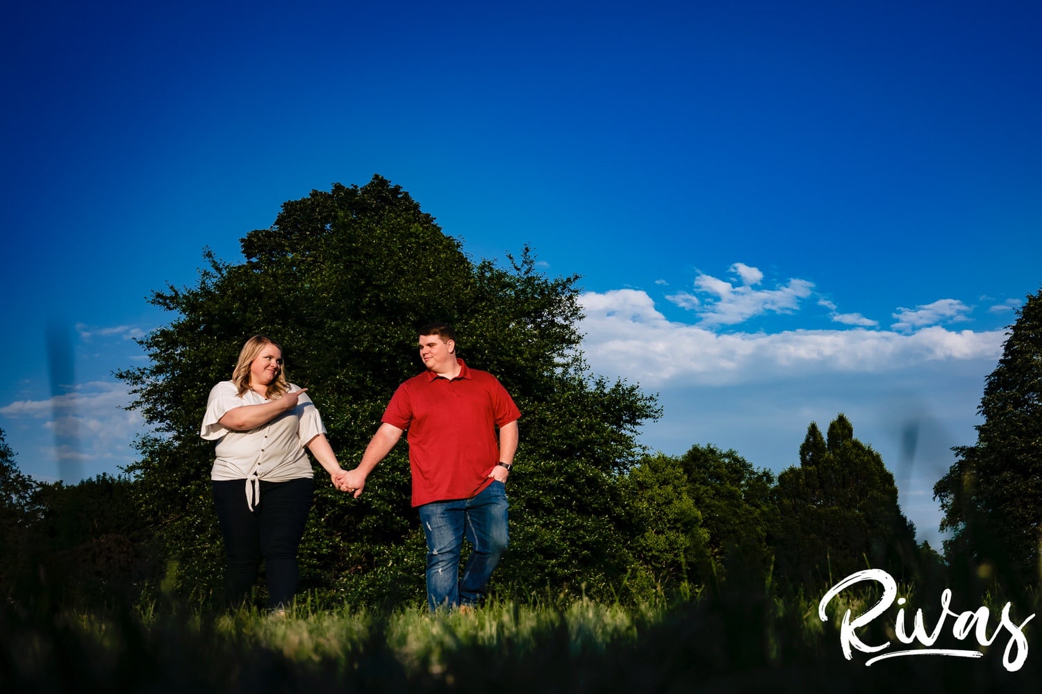 A wide, colorful picture taken from a distance of an engaged couple holding hands as they walk across the lawn at Loose Park during their engagement photography session in Kansas City. 