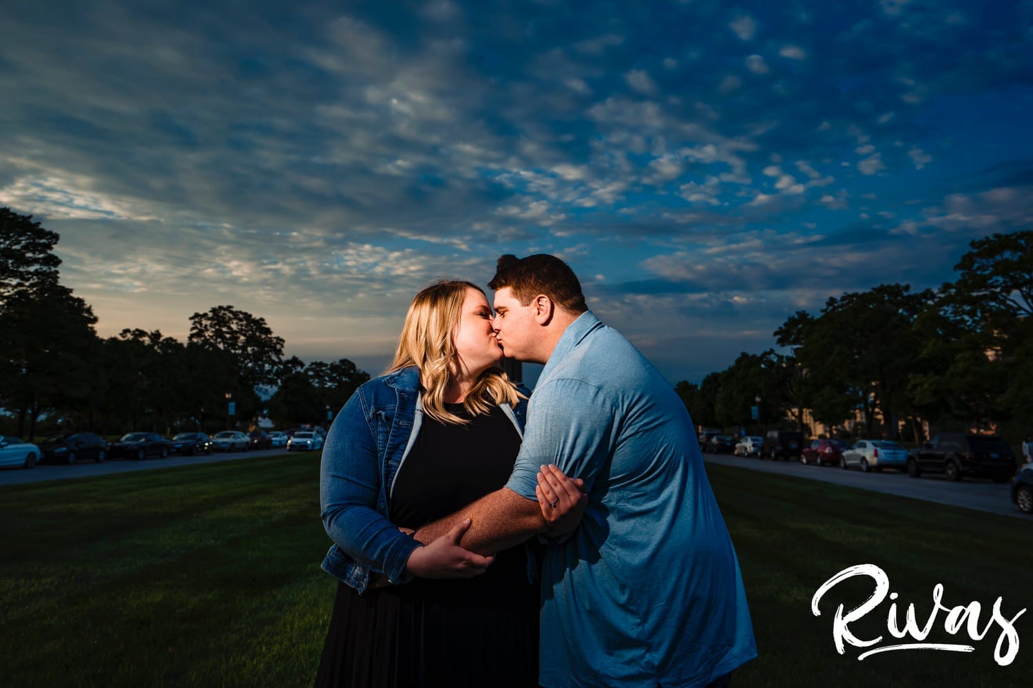 A colorful portrait of an engaged couple sharing an embrace and kiss in front of a bright blue sky with swirly clouds during their engagement photography session at Liberty Memorial in Kansas City. 