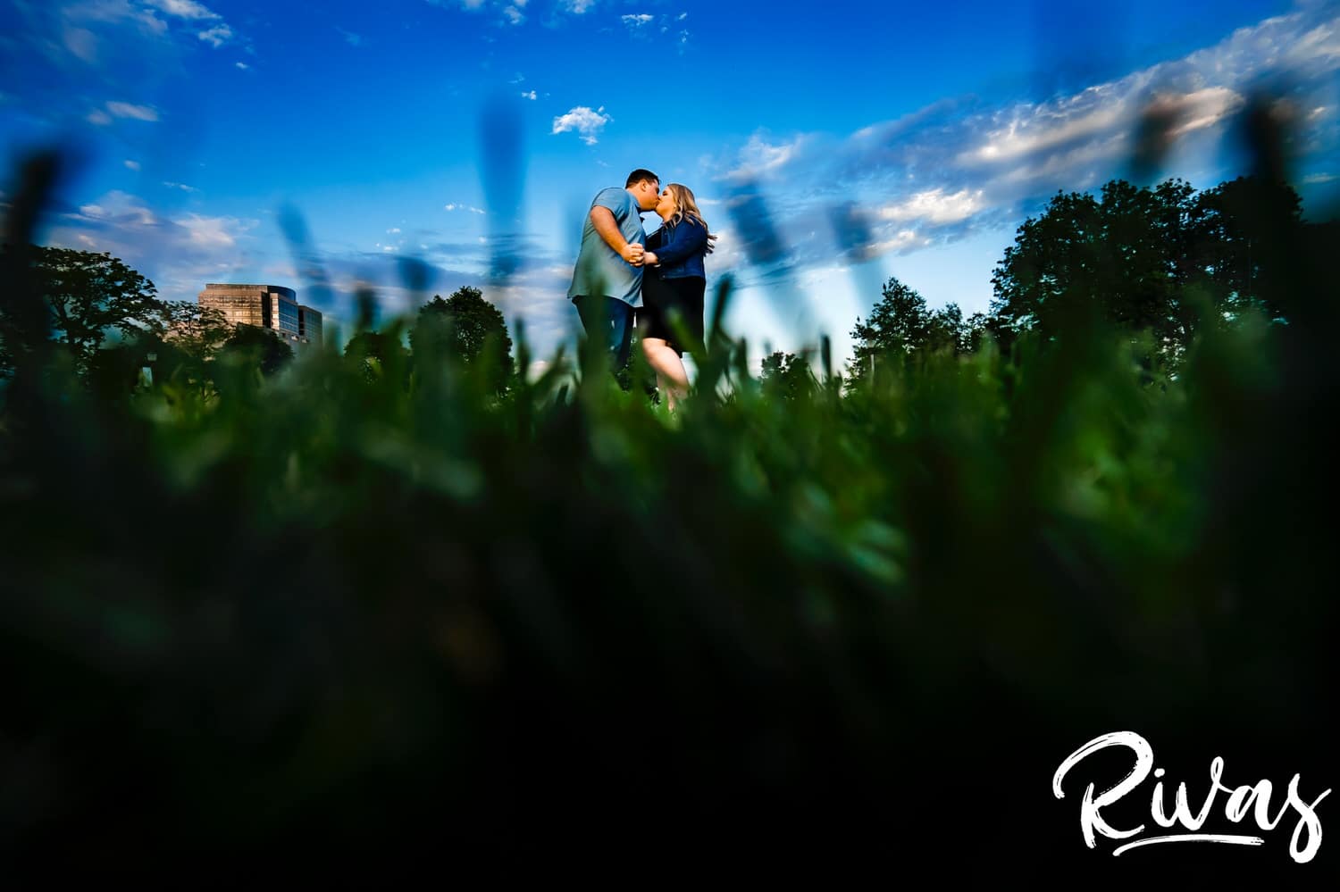 A wide, vibrant photograph taken from the ground up of an engaged couple sharing a kiss and embrace against a bright blue sky during their engagement photography session in Kansas City at Liberty Memorial. 