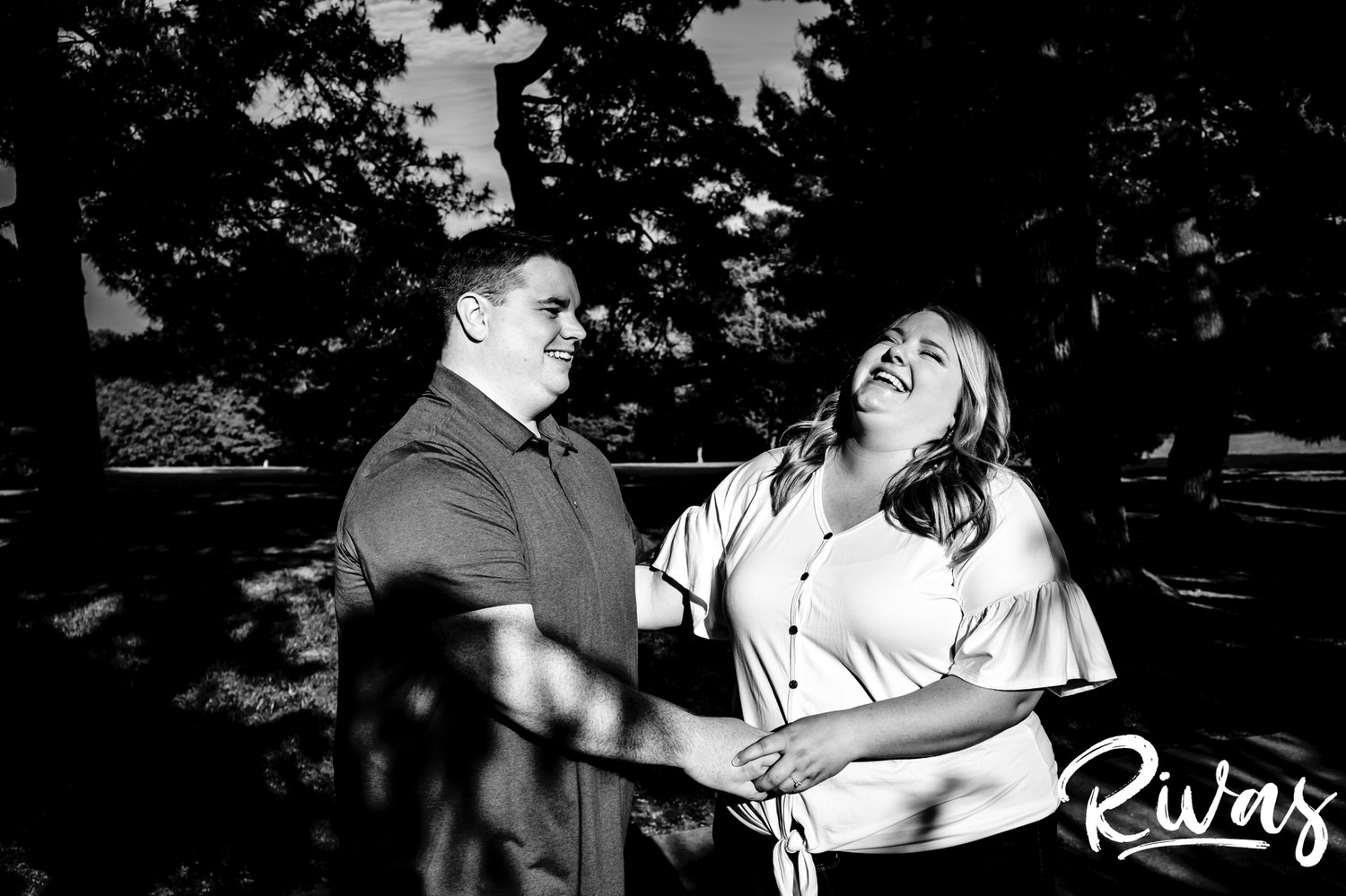 A candid, silly, black and white picture of an engaged couple laughing in a splash of sunlight underneath a canopy of dark trees during their engagement photography session at Loose Park in Kansas City. 