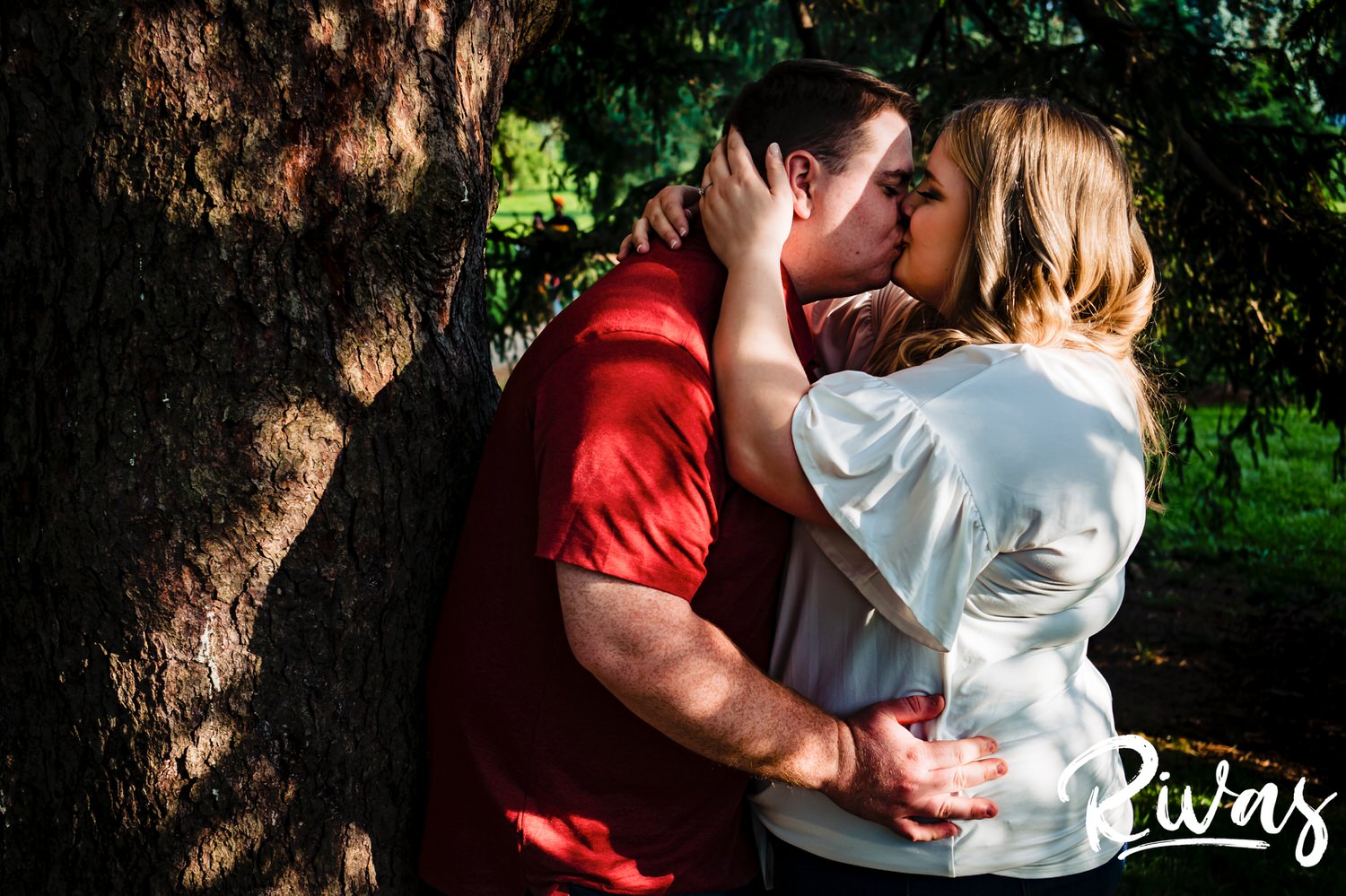 A close-up, candid picture of an engaged couple standing underneath a tree sharing an embrace and kiss during their summer engagement photography session at Kansas City's Loose Park