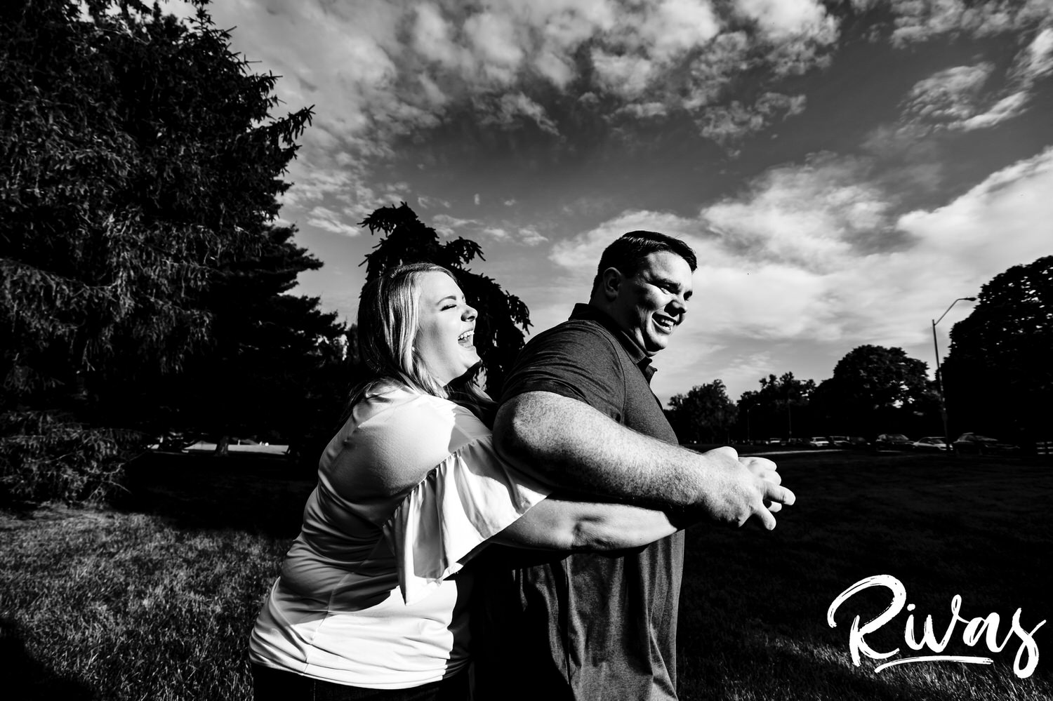 A close-up, fun, candid black and white picture of a woman sneaking up behind her fiance and tickling him as he laughs and smlies during their engagement photography session at Loose Park in Kansas City. 