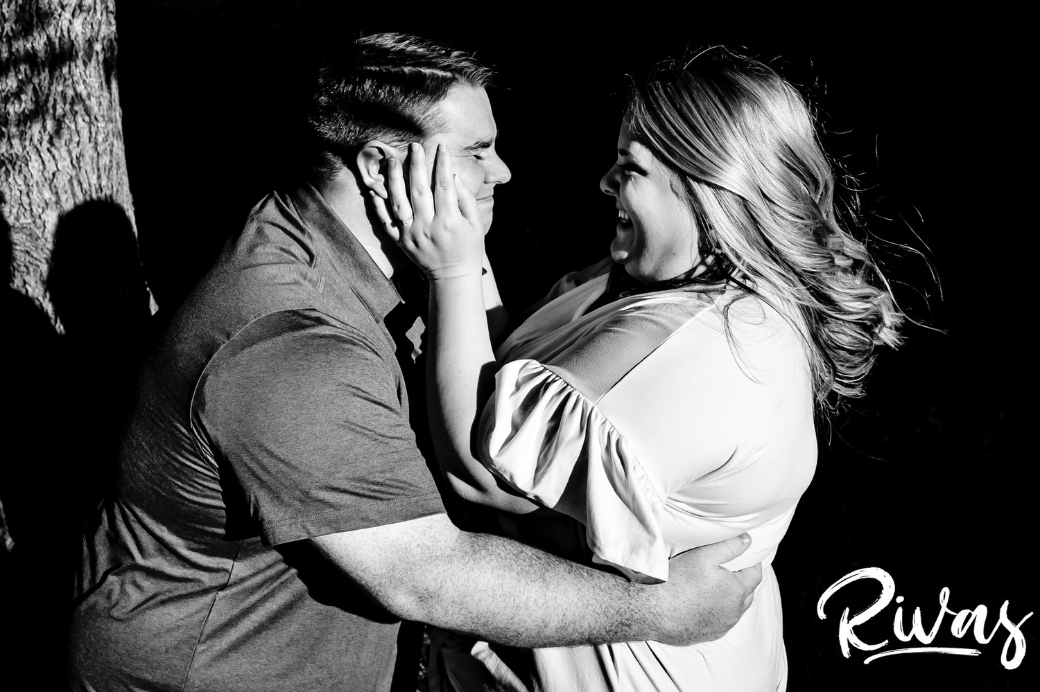 A candid black and white picture of a man tickling his fiance as she cradles his face in her hands during their summer engagement photography session in Kansas City at Loose Park. 
