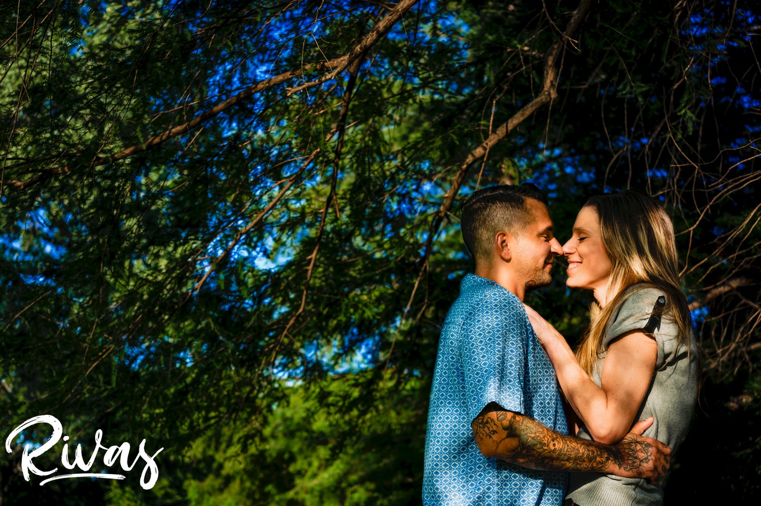 A colorful, intimate picture of an engaged couple sharing an embrace as they stand underneath a canopy of green trees during their summer engagement session at Shawnee Mission Park in Kansas City. 