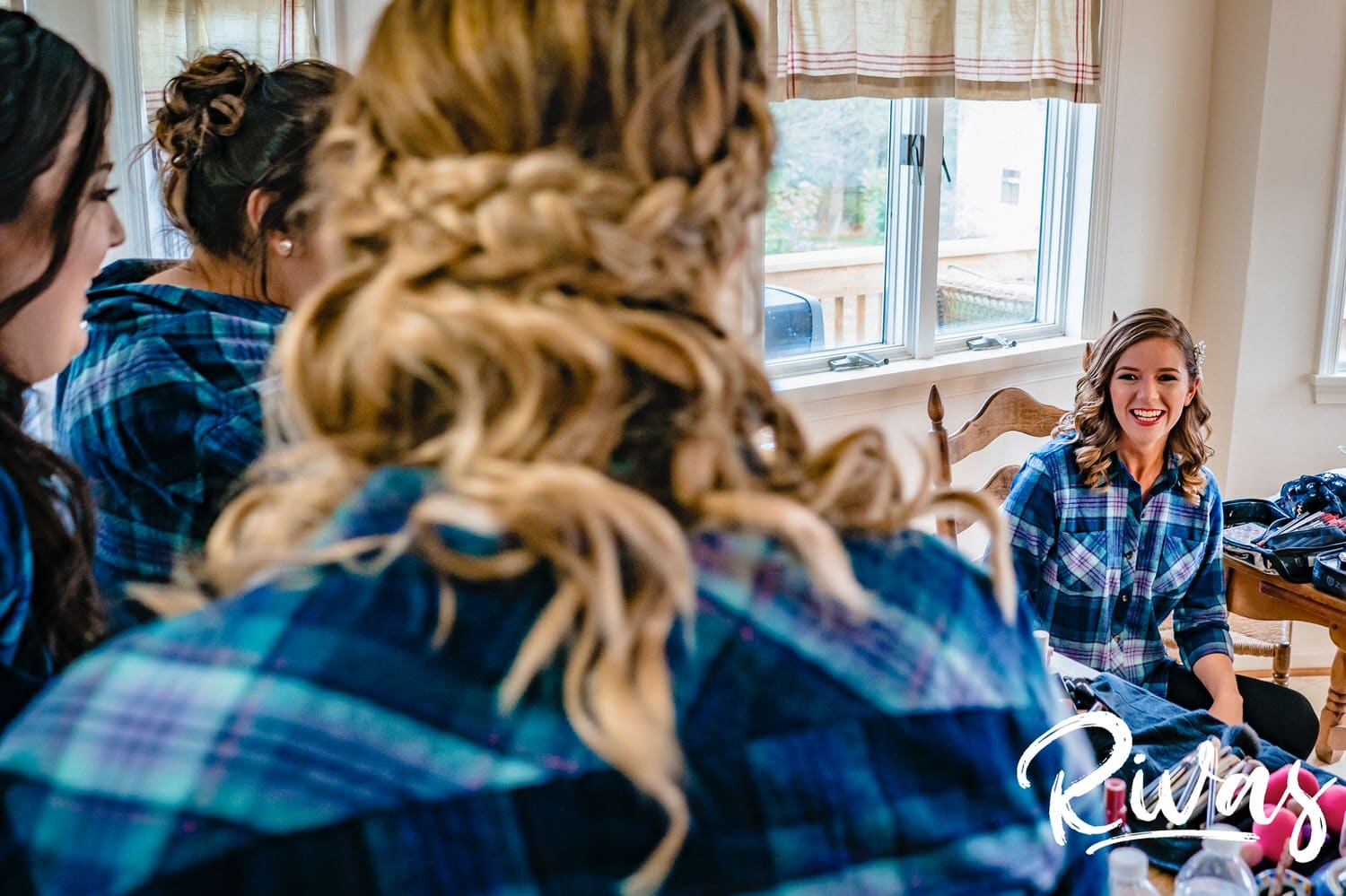 A colorful, candid picture taken over the shoulder of a bridesmaid in a flannel shirt of a bride smiling at her group of bridesmaids just after she's finished getting her makeup done on her fall wedding day at The Barns at Hamilton Station Vineyard. 