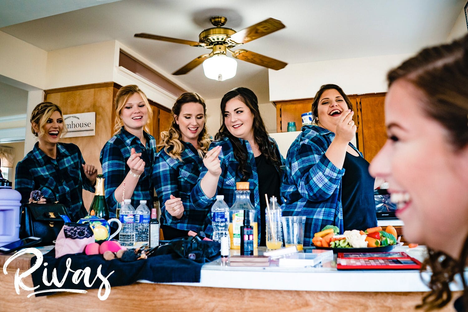 A candid picture taken over a bride's shoulder of  a group of bridesmaids in matching flannel shirts serenading her as she gets her makeup done on the afternoon of her Hamilton Station Vineyard fall wedding. 