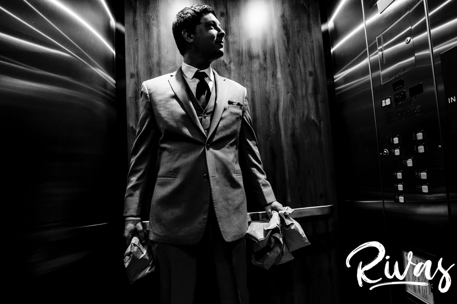A candid black and white picture of a groom in a gray tuxedo standing in an elevator clutching several brown paper bags on the afternoon of his fall wedding at Hamilton Station Vineyard. 
