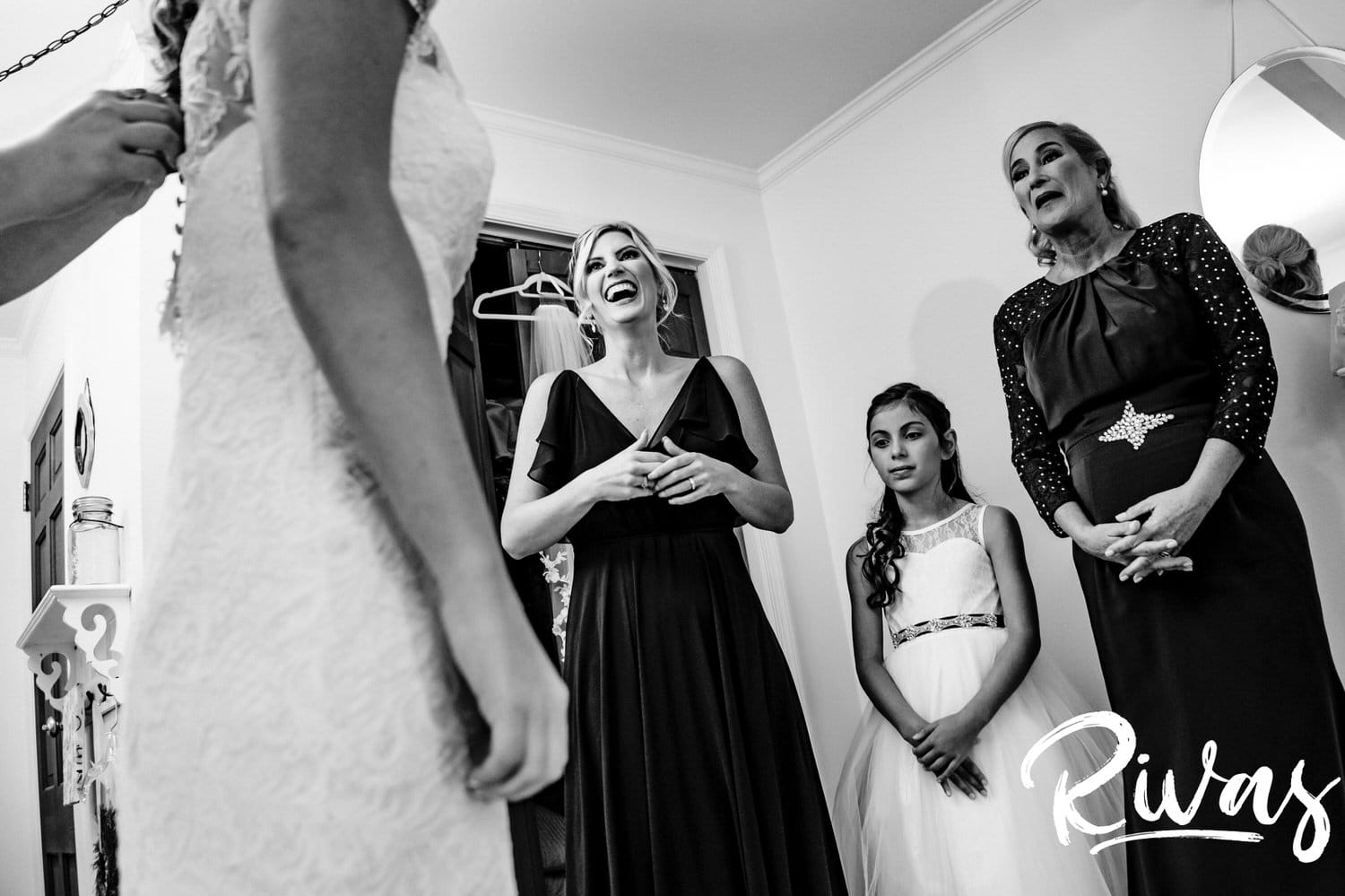 A candid black and white picture taken from behind a bride putting her gown on as her mom, sister, and niece watch on in adoration on the afternoon of her fall wedding at The Barns at Hamilton Station Vineyard. 