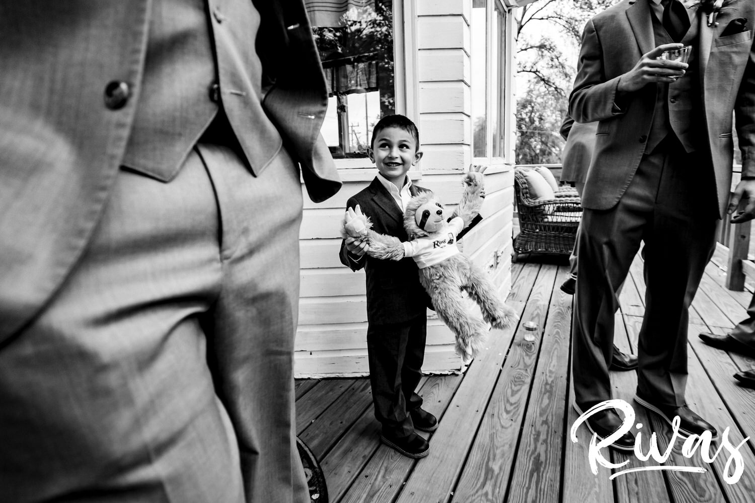 A candid black and white picture of a small ring bearer holding a stuffed sloth as he stands with the groom and groomsmen on a deck at The Barns at Hamilton Station Vineyard. 