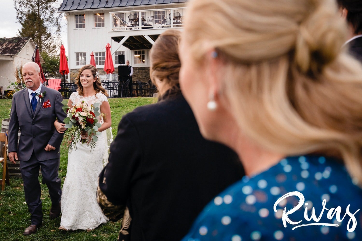 A colorful, candid picture taken over the shoulder of the bride's mom as the bride and her father walk down the aisle during her fall wedding ceremony at The Barns at Hamilton Station Vineyard. 