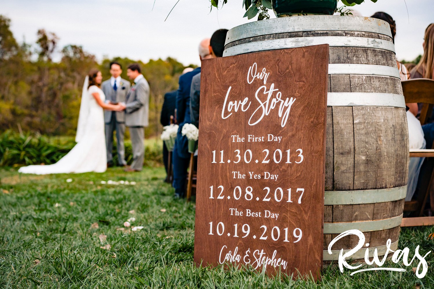 A picture of a couple's special dates on a wood sign at the back of a wedding aisle with the bride and groom visible at the front of the aisle. 