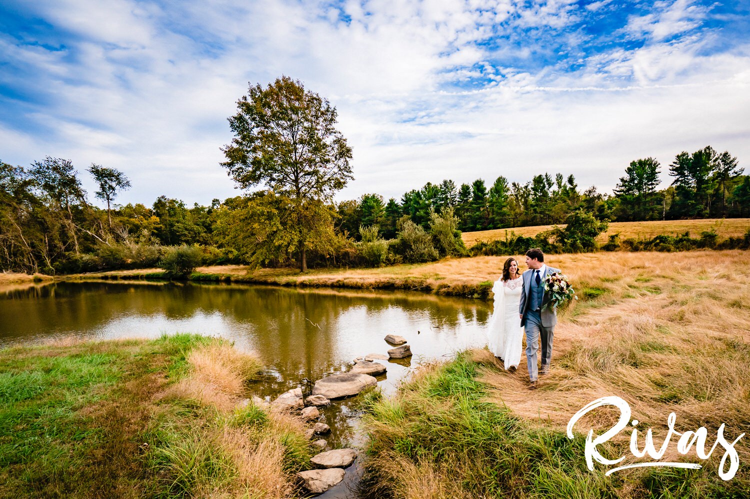 A colorful, wide picture of a bride and groom holding hands walking across a large field on their wedding day at The Barns at Hamilton Station Vineyard. 