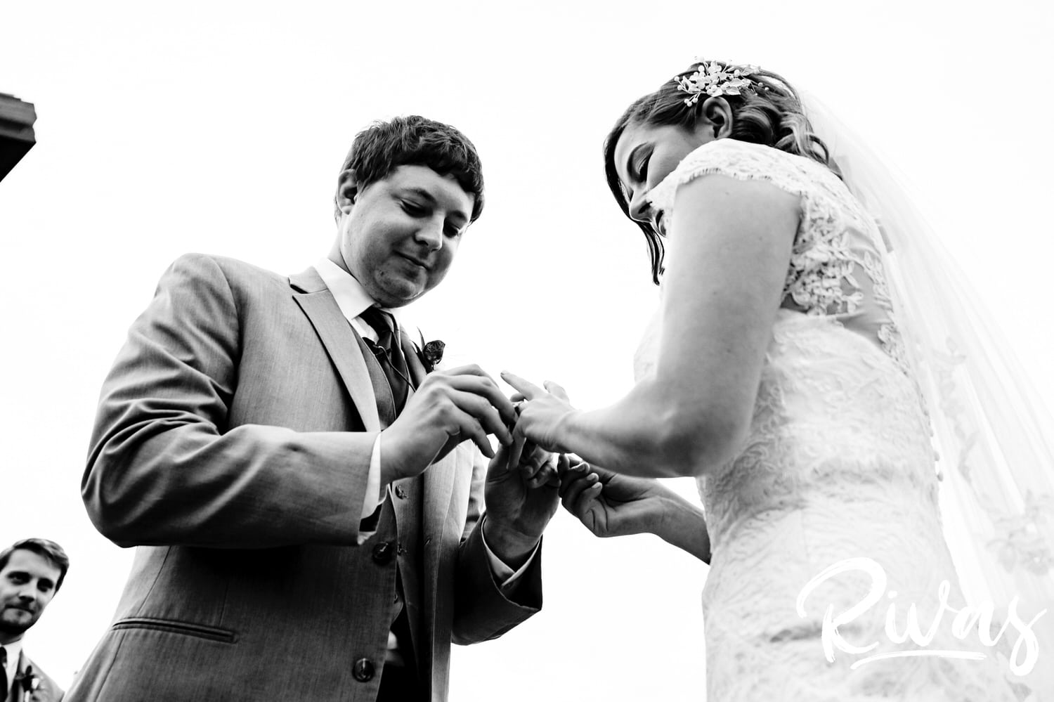 A black and white picture taken from below of a groom putting a wedding band on his bride's finger during their fall wedding ceremony. 